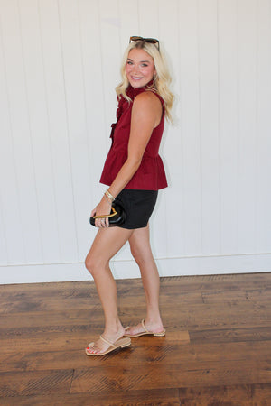 side view of young woman modeling a Texas A&M game day outfit - Lani Top with sleeveless burgundy peplum blouse, black shorts, and beige sandals